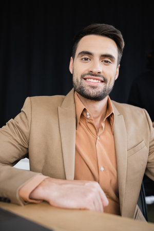 bearded businessman in beige blazer smiling at camera while sitting in officeの写真素材