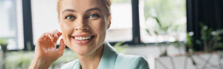 portrait of cheerful businesswoman holding hand near face and smiling at camera in office, banner