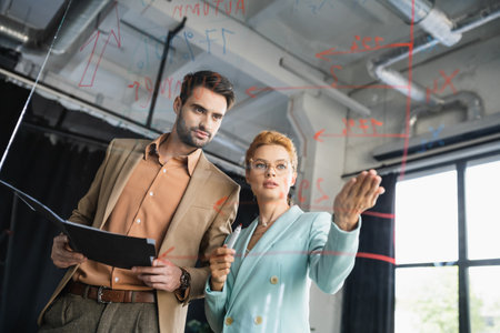 redhead businesswoman pointing at lettering on glass board near manager with folder in office