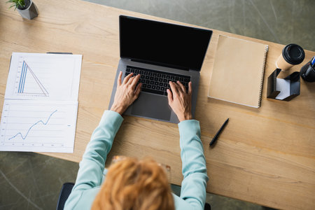 top view of redhead businesswoman working on laptop near papers with graphs in officeの写真素材