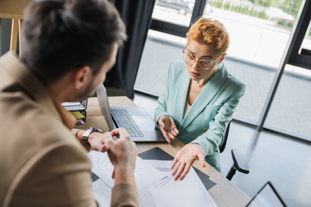 frowning businesswoman in eyeglasses pointing at analytics near blurred colleague in officeの写真素材