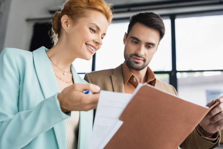 cheerful redhead businesswoman showing documents to bearded colleague in officeの写真素材