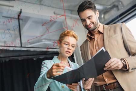 businesswoman in eyeglasses looking at documents near smiling colleague and glass board in officeの写真素材