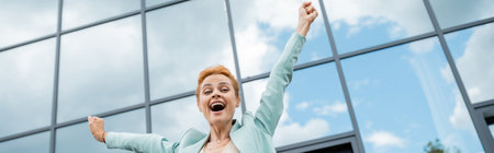 low angle view of cheerful redhead businesswoman celebrating triumph near modern building in city, bannerの写真素材