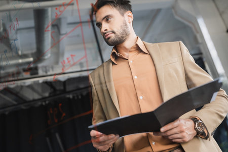 low angle view of bearded businessman with folder thinking near glass board with lettering in officeの写真素材