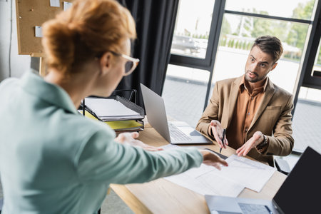 irritated business colleagues pointing at documents near laptops while quarreling in office