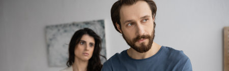 Dissatisfied bearded man looking away near blurred girlfriend at home, banner