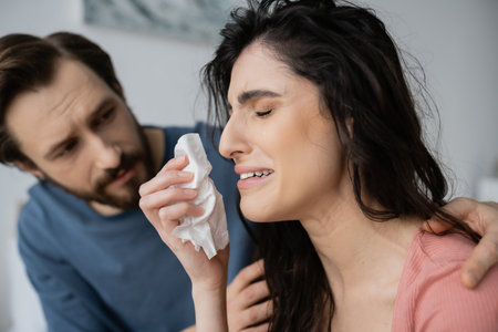 Blurred man calming down displeased and crying girlfriend with napkin at homeの写真素材