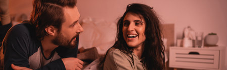Cheerful brunette woman talking to bearded boyfriend in bedroom at night, banner