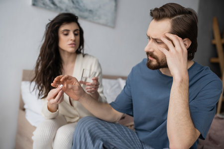 Man with headache taking pill from blurred caring girlfriend in bedroomの写真素材