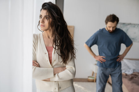 Displeased woman standing near curtains during relationship crisis with blurred boyfriend at homeの写真素材