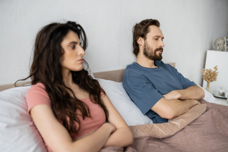 Dissatisfied man crossing arms while sitting near blurred girlfriend on bed at homeの写真素材