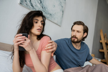 Jealous man looking at brunette girlfriend hiding smartphone in bedroom at homeの写真素材