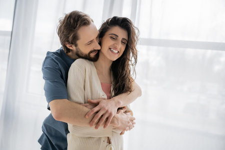 Bearded man embracing cheerful brunette girlfriend at homeの写真素材