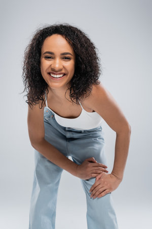 excited african american woman with curly brunette hair looking at camera isolated on greyの写真素材