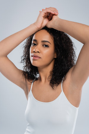 portrait of young african american woman with curly brunette hair posing with hands above head isolated on greyの写真素材
