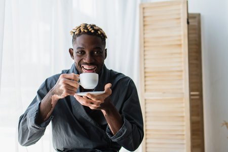 Happy african american man with vitiligo holding coffee and looking at camera at homeの写真素材