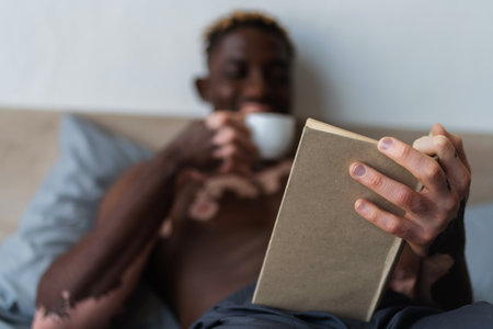 Blurred african american man with vitiligo holding cup and reading book in bedの写真素材
