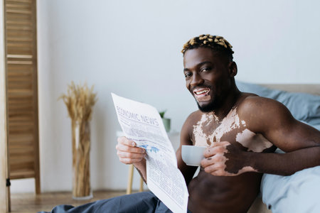 Cheerful african american man with vitiligo looking at camera and holding coffee with newspaper in bedroomの写真素材