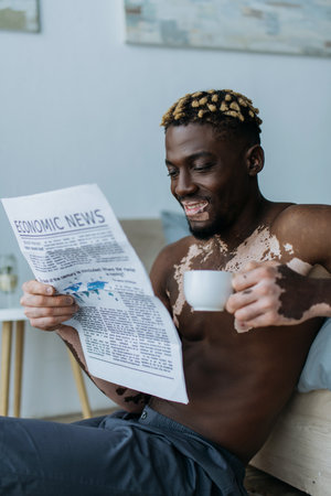Smiling african american man with vitiligo reading economic newspaper and holding coffee in bedroomの写真素材