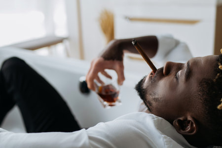 Side view of african american man with vitiligo smoking cigar and holding blurred whiskey in bathtubの写真素材