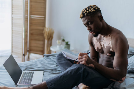 Shirtless african american freelancer with vitiligo writing on notebook near laptop on bedの写真素材