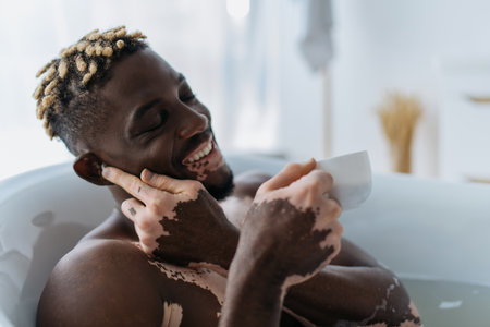 Overjoyed african american man with vitiligo using wireless earphone and holding coffee cup in bathtub at homeの写真素材