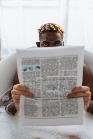 African american man with vitiligo covering face with newspaper in bath with foam at home