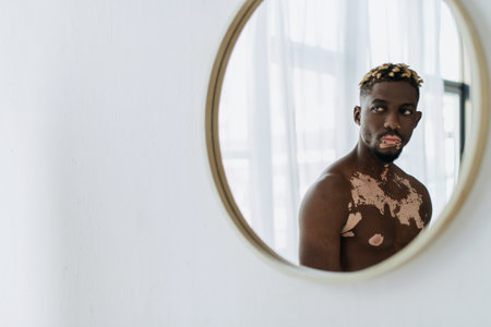 Shirtless african american man with vitiligo reflecting in mirror of modern bathroomの写真素材