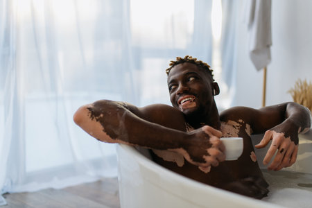 Cheerful african american man with vitiligo in earphone holding coffee cup and taking bath at homeの写真素材
