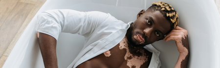 Top view of african american man with vitiligo in shirt lying in bathtub at home, bannerの写真素材