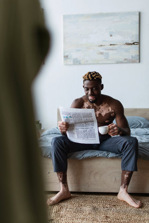 Smiling african american man with vitiligo reading news and holding coffee on bed at homeの写真素材