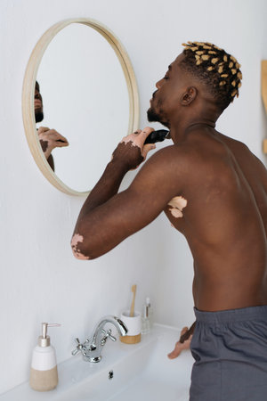 Side view of african american man with vitiligo shaving with electric razor in bathroomの写真素材