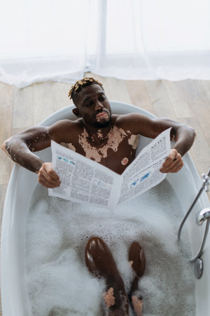 Top view of african american man with vitiligo reading newspaper while taking bath at homeの写真素材