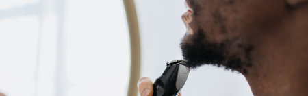 Cropped view of african american man with vitiligo shaving beard with electric razor in bathroom, bannerの写真素材
