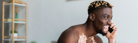 Smiling and shirtless african american man with vitiligo talking on smartphone at home, bannerの写真素材