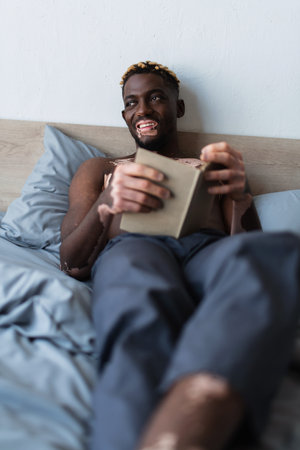 Shirtless and smiling african american man with vitiligo holding book while lying on bed at homeの写真素材