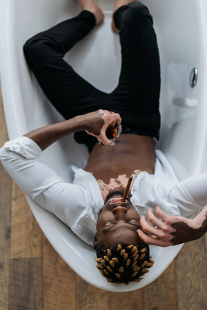 Overhead view of african american man with vitiligo in clothes smoking cigar and holding whiskey in...