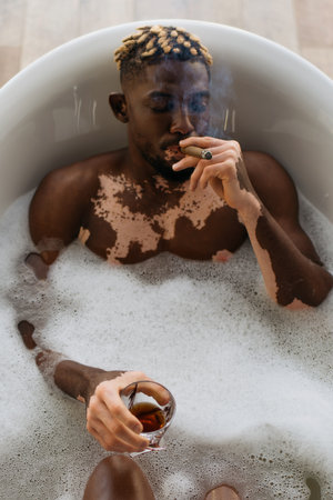 Top view of african american man with vitiligo smoking cigar and holding whiskey in bath with foamの写真素材