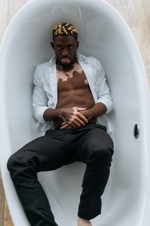 Top view of african american man with vitiligo in shirt and pants lying in bathtub at homeの写真素材