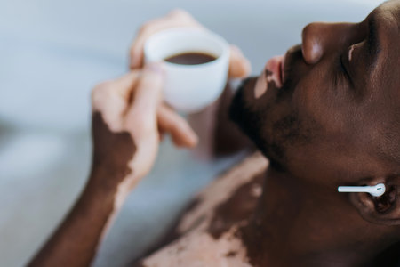 African american man with vitiligo using earphone and holding blurred coffee while taking bath at homeの写真素材