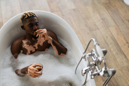 Top view of young african american man with vitiligo smoking cigar and holding glass of bourbon in bath with foamの写真素材