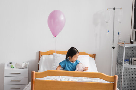little asian girl playing with toy bunny on bed under festive balloon in hospital wardの写真素材