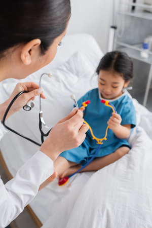 young doctor near blurred asian girl holding toy stethoscope while sitting on hospital bedの写真素材