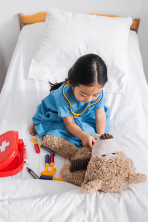 high angle view of asian girl examining teddy bear with toy stethoscope while playing on bed in hospitalの写真素材