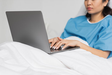 partial view of asian woman typing on laptop on bed in hospital wardの写真素材