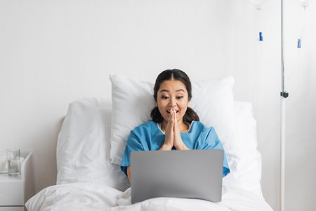 excited asian woman gesturing during video call on laptop in hospital wardの写真素材