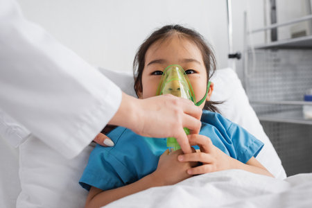little asian girl breathing in oxygen mask near doctor in hospital wardの写真素材