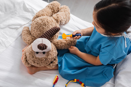 top view of asian kid in hospital gown holding toy reflex hammer near teddy bear while playing in clinicの写真素材