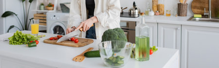 Cropped view of young woman cutting cherry tomatoes near vegetables and orange juice in kitchen,...